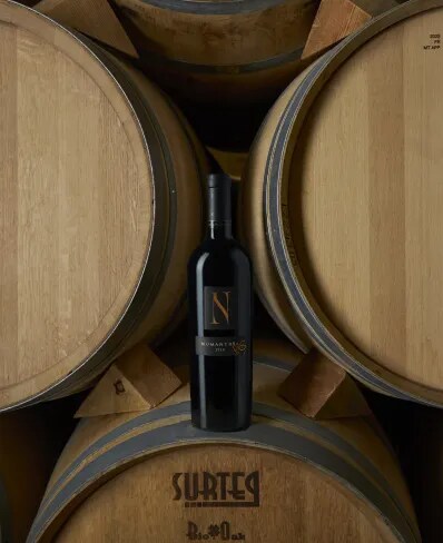 Botella de vino tinto español Numanthia 2018 apoyada contra barricas de roble tradicionales en Bodega Numanthia, destacando el meticuloso proceso de envejecimiento detrás de este vino premium.