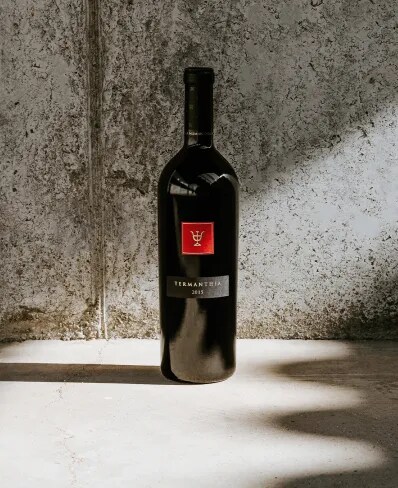 Vino tinto español Termanthia 2015 de Bodega Numanthia, presentado en un entorno minimalista con paredes de concreto texturizado y una iluminación dramática que proyecta sombras en el suelo.