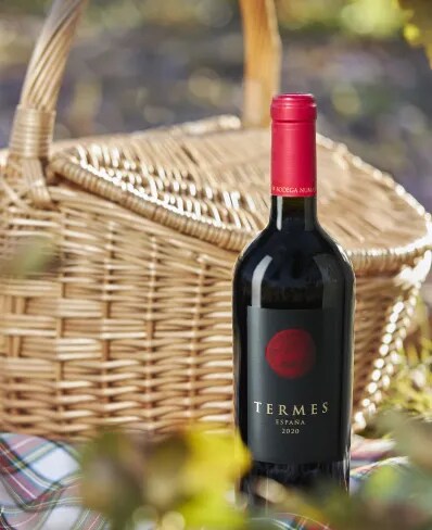Botella de vino tinto español Termes 2020 de Bodega Numanthia, expuesta al aire libre junto a una cesta de picnic de mimbre sobre un mantel a cuadros, rodeada de naturaleza.